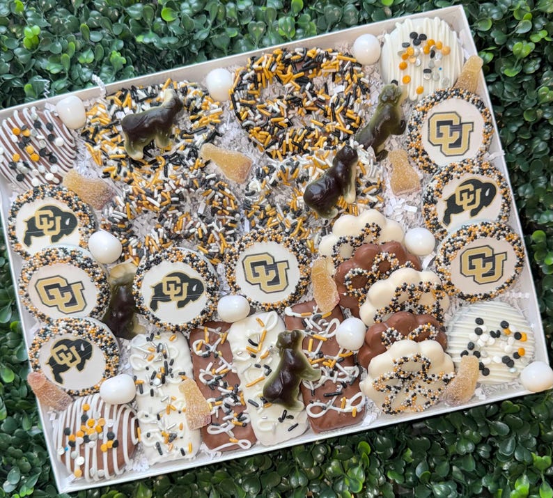 May include: A white rectangular box filled with an assortment of decorated treats. The treats include cookies with a university logo, pretzels with sprinkles, and chocolate-covered items. The color scheme is black, gold, and white.