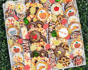 Puede incluir: Una caja blanca llena de una variedad de golosinas cubiertas de chocolate. Las golosinas incluyen pretzels, galletas y barras, decoradas con chispas, frutas y texto. La caja está sobre un fondo verde.