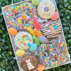May include: A white box filled with Easter treats. The box contains chocolate-covered biscuits and pretzels decorated with colourful sprinkles, a bunny, and pastel-coloured sweets. The treats are arranged on shredded paper.