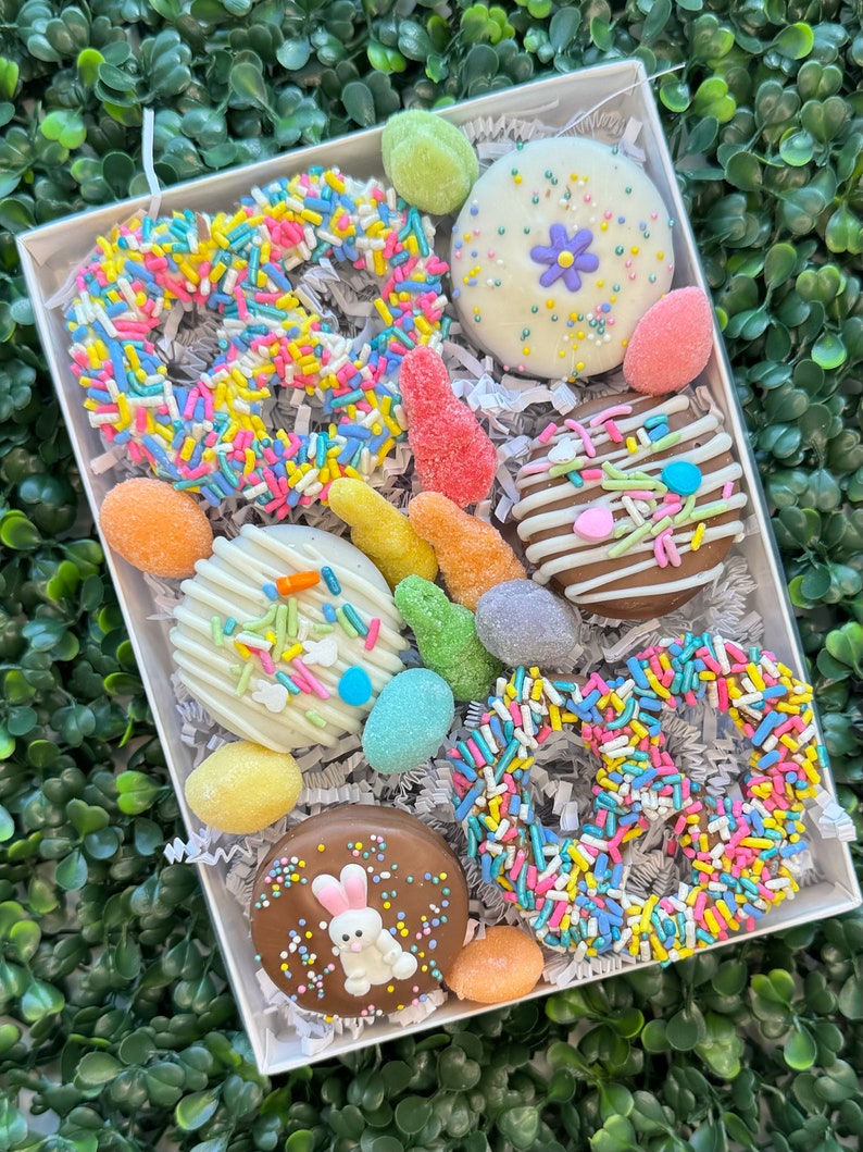 May include: A white box filled with Easter treats. The box contains chocolate-covered biscuits and pretzels decorated with colourful sprinkles, a bunny, and pastel-coloured sweets. The treats are arranged on shredded paper.