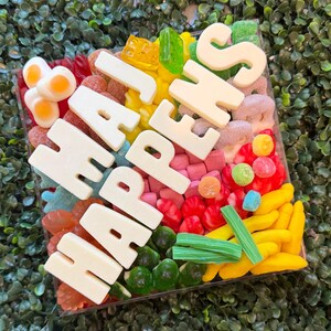 May include: A square clear container filled with a variety of colorful gummy candies, including yellow bananas, red berries, green bricks, and fried egg shapes. Large white chocolate letters spell out the phrase HAJ HAPPENS across the top.