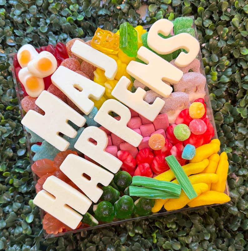 May include: A square clear container filled with a variety of colorful gummy candies, including yellow bananas, red berries, green bricks, and fried egg shapes. Large white chocolate letters spell out the phrase HAJ HAPPENS across the top.