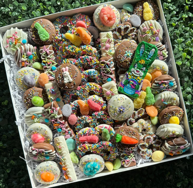 May include: A white box filled with Easter confectionery. Chocolate-covered pretzels, biscuits, and sweets are adorned with pastel sprinkles, bunny and carrot motifs. A green foil-wrapped chocolate bunny is among the treats.