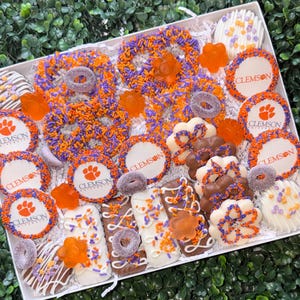 May include: A gift box filled with assorted treats in orange, purple, and white. The box includes pretzels, cookies, and chocolate-covered items, many with the Clemson paw print logo. The treats are arranged on a white surface.