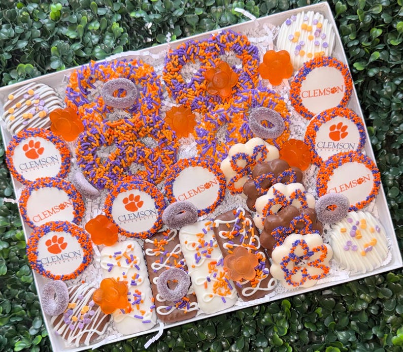 May include: A gift box filled with assorted treats in orange, purple, and white. The box includes pretzels, cookies, and chocolate-covered items, many with the Clemson paw print logo. The treats are arranged on a white surface.
