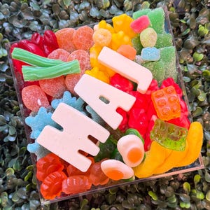 May include: A square clear container filled with a variety of colorful gummy candies, including red, orange, yellow, green, and blue shapes. Large white gummy letters spelling out the word HAJ are placed prominently across the center of the mix.