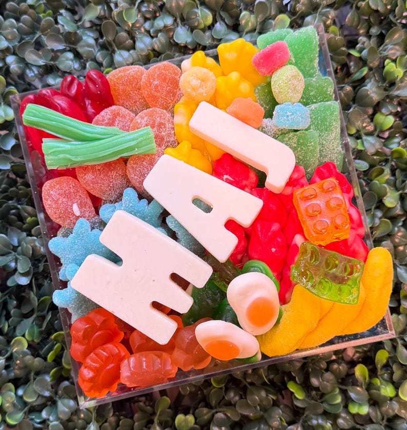 May include: A square clear container filled with a variety of colorful gummy candies, including red, orange, yellow, green, and blue shapes. Large white gummy letters spelling out the word HAJ are placed prominently across the center of the mix.