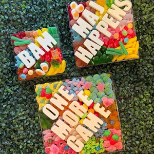 May include: Three square clear containers filled with assorted colorful gummy candies. The top left container features white letters spelling BAM. The top right container spells MAJ HAPPENS. The bottom container spells ONE MORE GAME.