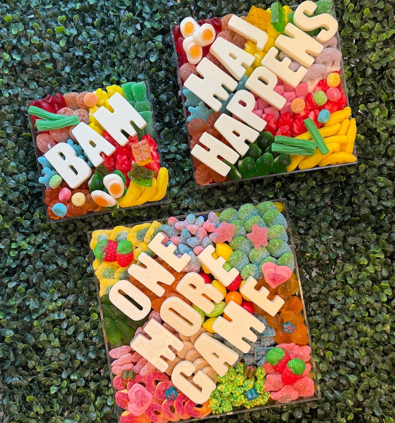 May include: Three square clear containers filled with assorted colorful gummy candies. The top left container features white letters spelling BAM. The top right container spells MAJ HAPPENS. The bottom container spells ONE MORE GAME.