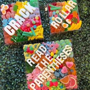 May include: Three square clear containers filled with assorted colorful gummy candies. The white lettering on the containers spells out CRACK, ROLLAR, and READ THE PARENTHESES. The candies include various shapes like bananas, eggs, and berries.