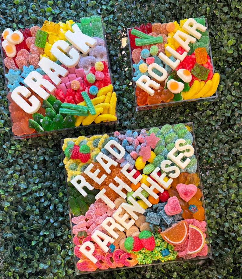 May include: Three square clear containers filled with assorted colorful gummy candies. The white lettering on the containers spells out CRACK, ROLLAR, and READ THE PARENTHESES. The candies include various shapes like bananas, eggs, and berries.