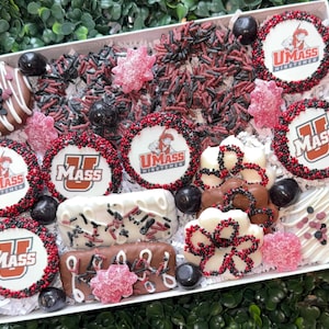 May include: A gift box filled with assorted decorated cookies and candies. The cookies feature the UMass logo in red and white, surrounded by red and black sprinkles. Other treats include chocolate-covered items and pink candies.