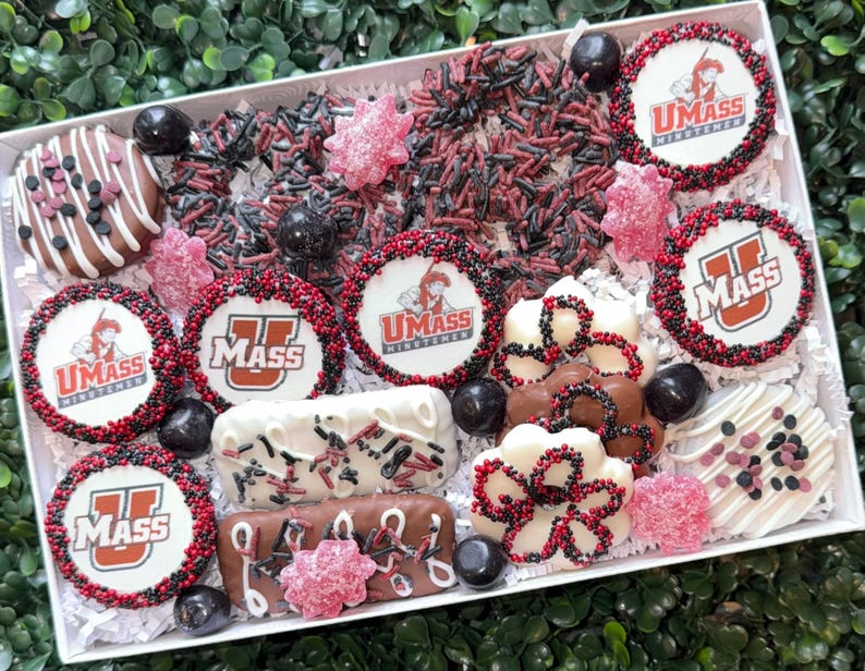 May include: A gift box filled with assorted decorated cookies and candies. The cookies feature the UMass logo in red and white, surrounded by red and black sprinkles. Other treats include chocolate-covered items and pink candies.