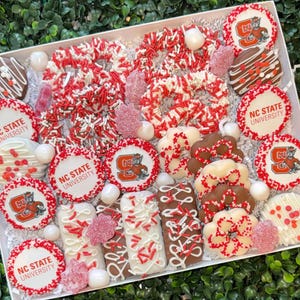 May include: A white box filled with assorted treats in red, white, and brown. The treats include cookies with the NC State University logo, chocolate-covered pretzels, and other sweets decorated with sprinkles and icing.