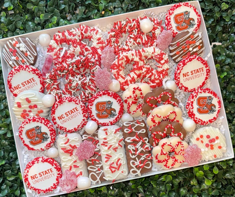 May include: A white box filled with assorted treats in red, white, and brown. The treats include cookies with the NC State University logo, chocolate-covered pretzels, and other sweets decorated with sprinkles and icing.
