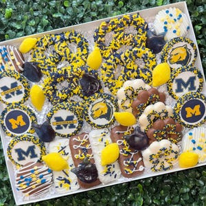 May include: A white box filled with an assortment of decorated treats. The treats include pretzels, cookies, and chocolate-covered items, all adorned with yellow and navy blue sprinkles and designs. Some treats feature the letter "M" or the word "Michigan".