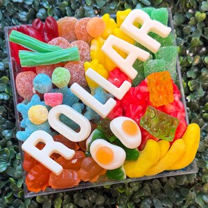 May include: A square clear container filled with a variety of colorful gummy candies, including green licorice, orange slices, blue stars, and fried egg shapes, featuring white chocolate letters spelling out the word DOLLAR.