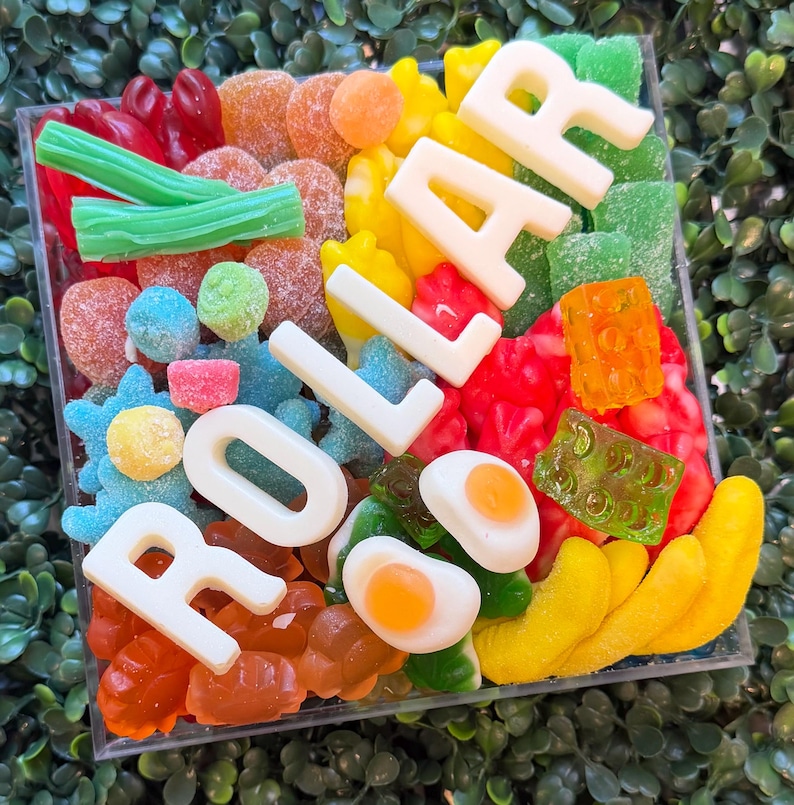 May include: A square clear container filled with a variety of colorful gummy candies, including green licorice, orange slices, blue stars, and fried egg shapes, featuring white chocolate letters spelling out the word DOLLAR.