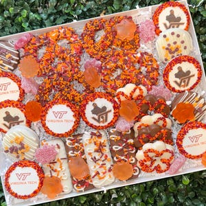 May include: A white box filled with an assortment of decorated cookies, pretzels, and candies. The cookies feature the Virginia Tech logo and a maroon and orange color scheme. The box is arranged on a bed of green foliage.