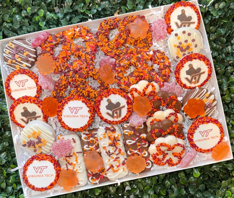 May include: A white box filled with an assortment of decorated cookies, pretzels, and candies. The cookies feature the Virginia Tech logo and a maroon and orange color scheme. The box is arranged on a bed of green foliage.