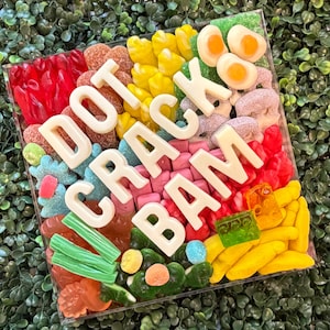 May include: A square clear container filled with an assortment of colorful gummy candies, including red, yellow, green, and pink varieties, topped with large white chocolate letters spelling out the phrase DOT CRACK BAM in a bold, block font.