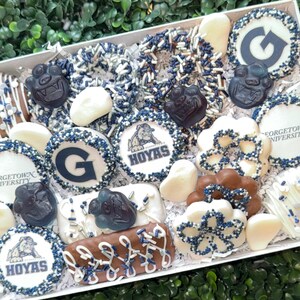 May include: A white box filled with assorted treats, including cookies, pretzels, and chocolates. The treats are decorated with blue and white sprinkles, and some feature the Georgetown University logo and the word "HOYAS".