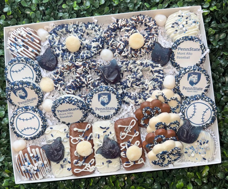 May include: A white box filled with assorted chocolate-covered treats. The treats include pretzels, cookies, and bars decorated with white, blue, and brown icing, sprinkles, and Penn State logos. Some treats are shaped like baseballs.