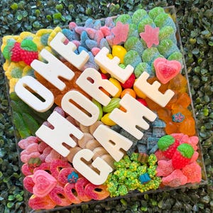 May include: A square clear acrylic box filled with a variety of colorful gummy candies, including strawberries, hearts, and rings. Large white block letters spelling out the phrase ONE MORE GAME are placed on top of the assorted candy mix.