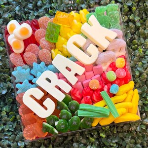 May include: A square clear container filled with an assortment of colorful gummy candies, including fried egg shapes, blue stars, red berries, and yellow bananas, featuring large white chocolate letters spelling out the word CRACK.