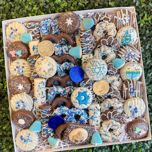 Könnte beinhalten: Eine weiße Box voller Hanukkah-Themen-Leckereien. Die Leckereien umfassen schokoladenüberzogene Brezeln, Kekse und Süßigkeiten, die mit blauen und weißen Streuseln, Sternen und Menora verziert sind. Einige Leckereien sind in weißer Schokolade getunkt.