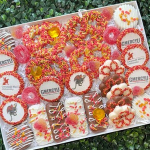 May include: A white box filled with an assortment of decorated cookies, pretzels, and candies. The cookies are round and decorated with red and yellow sprinkles, and the text "GMERCYU". Some cookies have a dragon logo.
