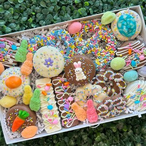 May include: A white box filled with an assortment of Easter-themed treats. The box contains chocolate-covered biscuits, decorated with sprinkles, pastel-coloured sweets, and a variety of shapes, including eggs, carrots, and a bunny.