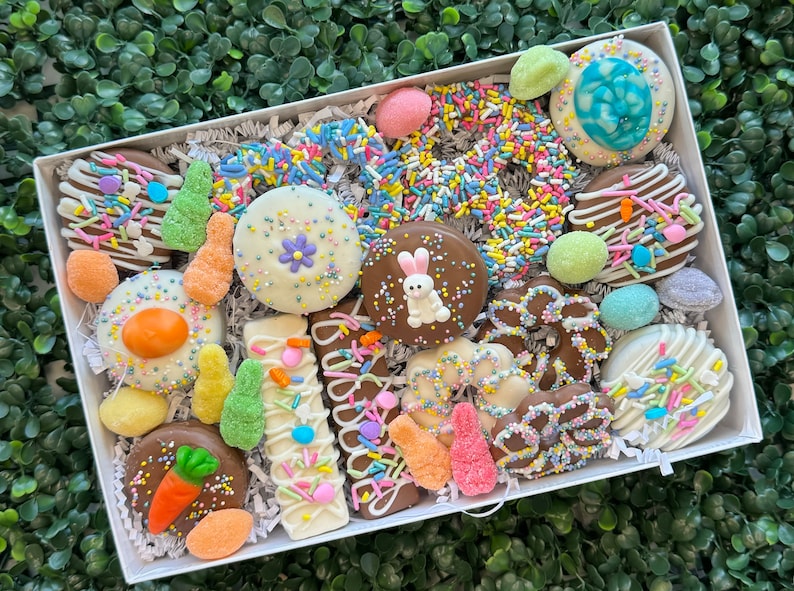 May include: A white box filled with an assortment of Easter-themed treats. The box contains chocolate-covered biscuits, decorated with sprinkles, pastel-coloured sweets, and a variety of shapes, including eggs, carrots, and a bunny.