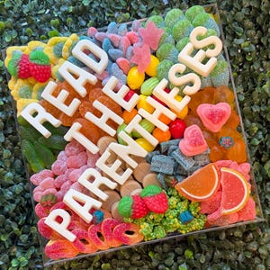May include: A square clear acrylic box filled with a variety of colorful gummy candies, including strawberries, orange slices, hearts, and rings. Large white block letters spelling READ THE PARENTHESES are arranged diagonally across the candy mix.