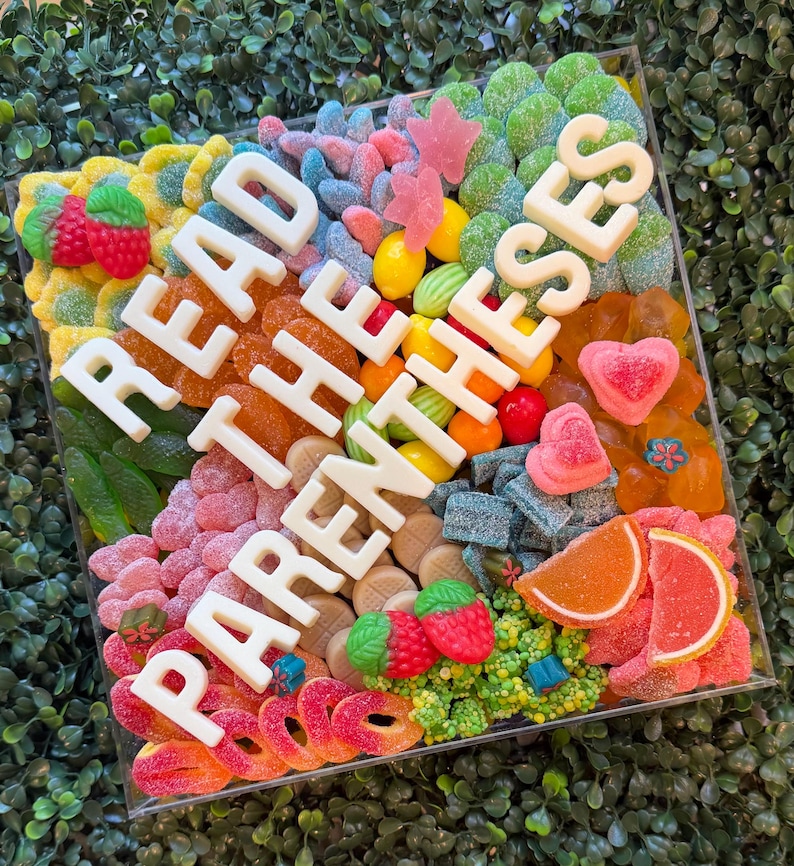 May include: A square clear acrylic box filled with a variety of colorful gummy candies, including strawberries, orange slices, hearts, and rings. Large white block letters spelling READ THE PARENTHESES are arranged diagonally across the candy mix.
