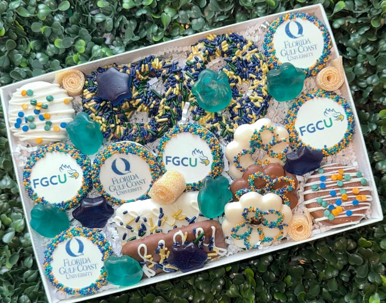 May include: A white gift box filled with assorted treats, including cookies, pretzels, and candies. The cookies feature the "FGCU" logo and the words "Florida Gulf Coast University". The treats are decorated with blue, yellow, and white sprinkles and accents.