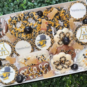 May include: A gift box filled with assorted treats, including decorated cookies, chocolate-covered items, and candies. The cookies feature the Appalachian logo and the letter "A" in gold and black. The treats are arranged on a white surface.