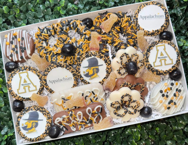 May include: A gift box filled with assorted treats, including decorated cookies, chocolate-covered items, and candies. The cookies feature the Appalachian logo and the letter "A" in gold and black. The treats are arranged on a white surface.