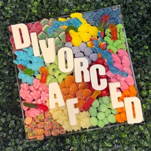 May include: A square clear tray filled with a variety of colorful gummy candies, including sour belts, rings, and fruit shapes. Large white block letters spelling out the word DIVORCED are arranged across the top of the candy assortment.