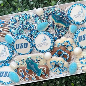 May include: A gift box filled with assorted treats, including cookies and candies, in shades of white, blue, and brown. Some cookies feature the "University of San Diego" logo and "USD" in blue. The box is decorated with sprinkles and candies.
