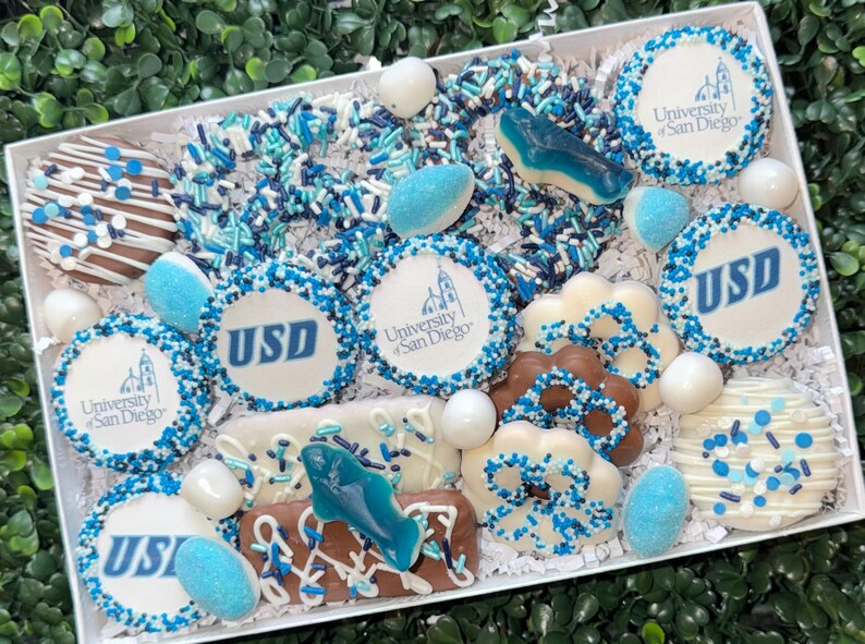 May include: A gift box filled with assorted treats, including cookies and candies, in shades of white, blue, and brown. Some cookies feature the "University of San Diego" logo and "USD" in blue. The box is decorated with sprinkles and candies.