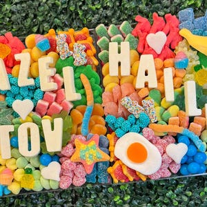 May include: A rectangular candy board featuring a colorful assortment of gummy candies, including frogs, bananas, hearts, and fried eggs. White chocolate letters spell out "MAZEL TOV" and "HAILEY" across the arrangement of sweets.