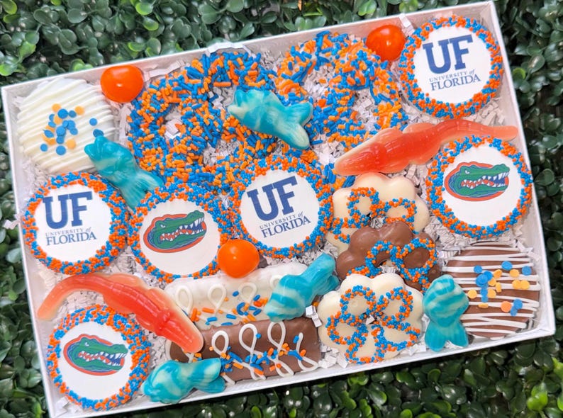 May include: A white box filled with assorted treats, including cookies with the University of Florida logo and Gator mascot, orange and blue sprinkles, and gummy candies. The treats are decorated in the school's colors.