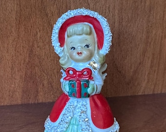 May include: A vintage Christmas figurine of an angel holding a gift. The angel wears a red dress and hat with white trim, and a green and red gift. The figurine has blonde hair and is made of ceramic.