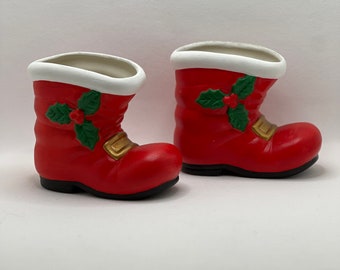 May include: A pair of red ceramic Santa boots with white fur trim and green holly accents. The boots are designed to be used as small planters or decorative items.
