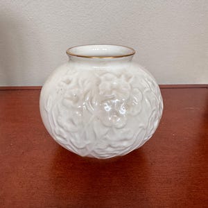 May include: A white ceramic vase with a gold rim and a raised floral design.