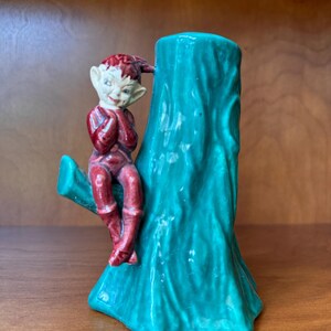 May include: A ceramic figurine of an elf sitting on a green tree stump. The elf is wearing a red outfit and has a white face.