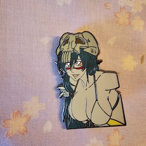 May include: Enamel pin featuring a stylized anime character with teal hair, red eye markings, and a skull headdress. The character wears a light beige bikini. The pin has a black outline and is set against a pink floral background.