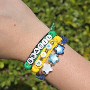 Könnte beinhalten: Ein Set aus drei gestapelten Armbändern mit einer grünen Kordel mit weißen Buchstabenperlen, die BRASIL buchstabieren, einer gelben Kordel mit kleinen Herzperlen und einer blauen Glitzerkordel mit drei sternförmigen Perlen in Weiß, Hellblau und Dunkelblau.
