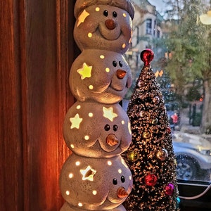 May include: A stack of five white ceramic snowmen with stars and lights, wearing a red Santa hat, next to a small, glittery, gold Christmas tree with red and gold ornaments.